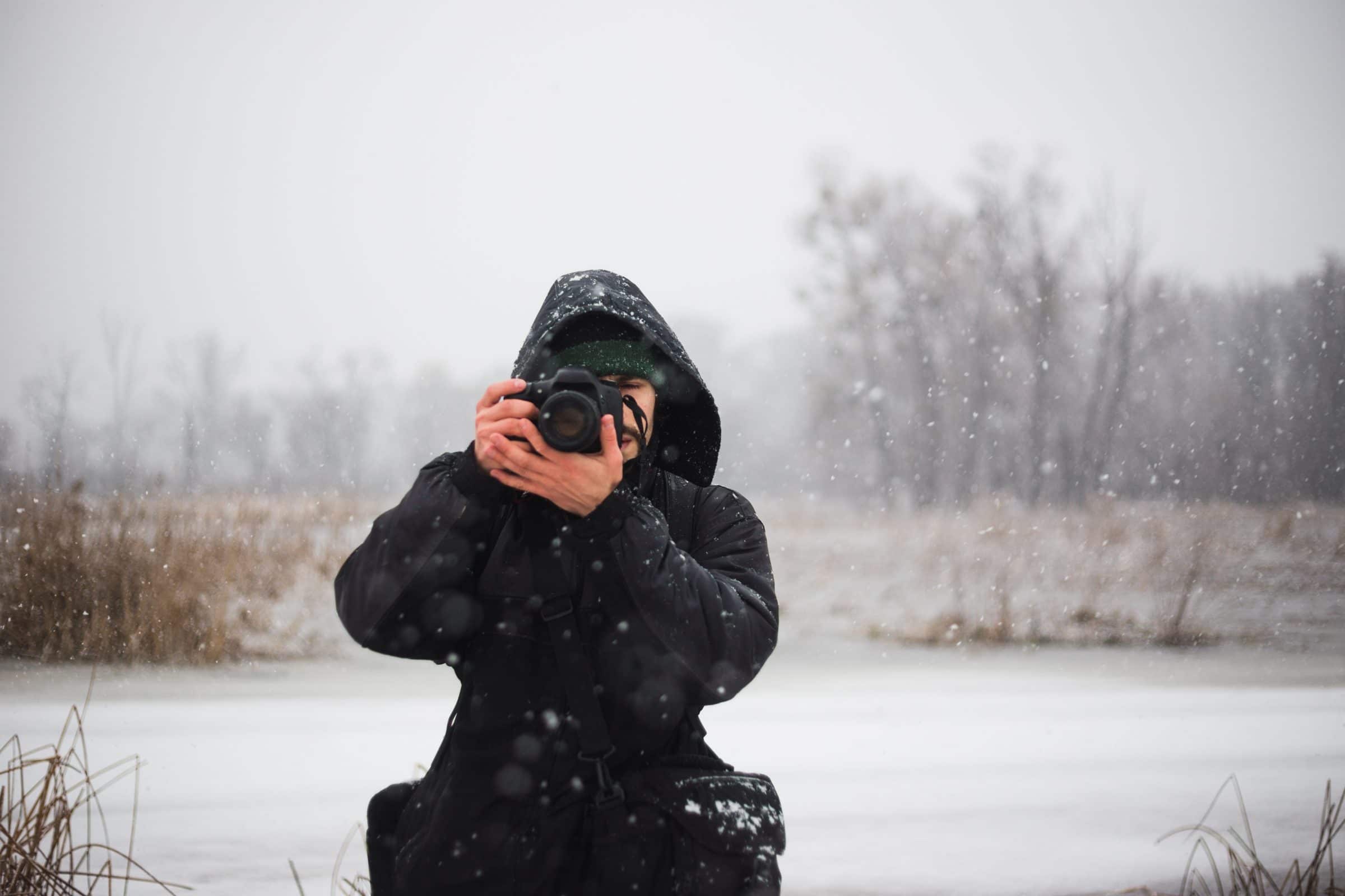 An adult stands in a winter field, the hood of their black winter jacket up. They hold a camera up to their eye and face the camera.