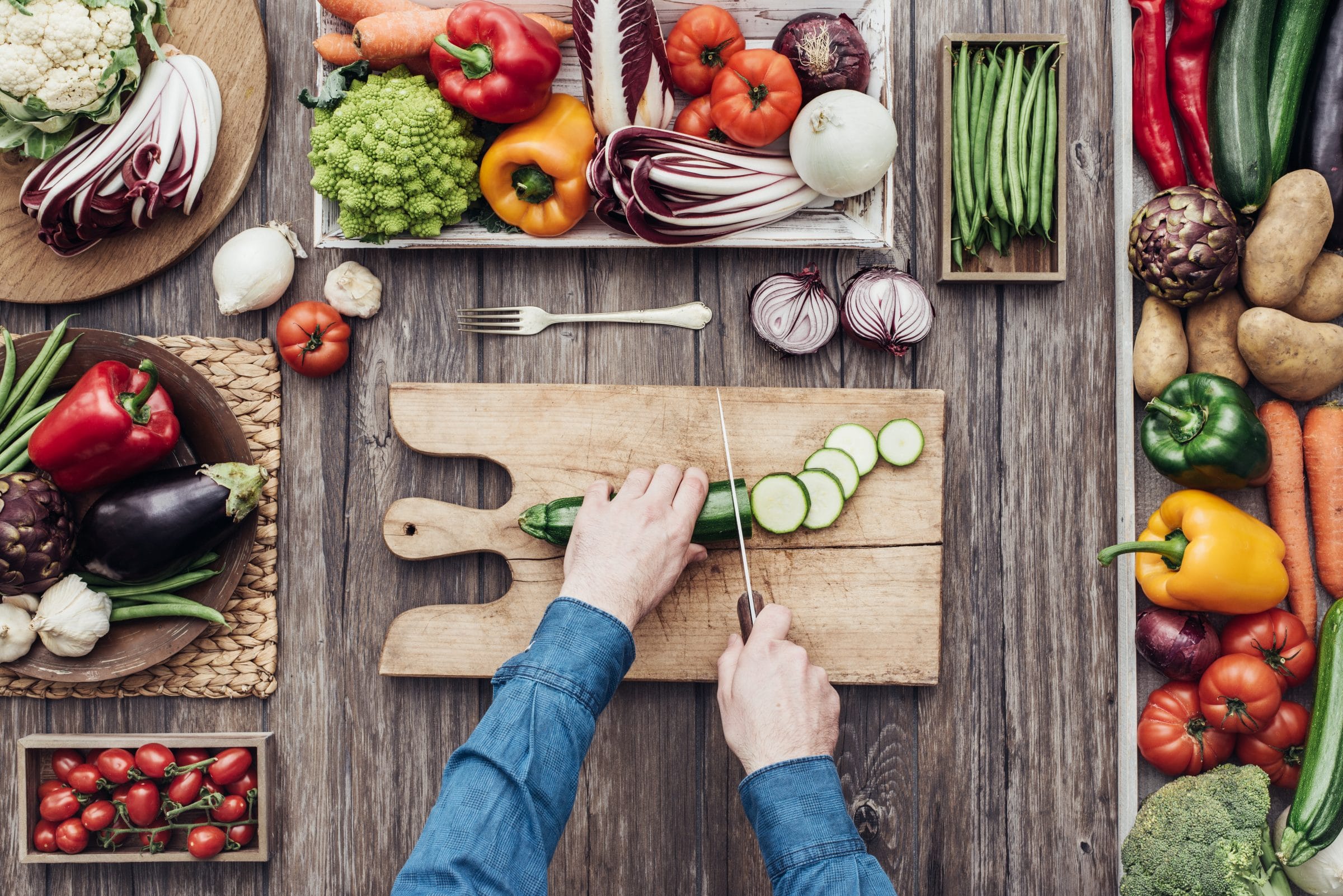 An overview shot of hands cutting a cucumber. Various colourful vegetables surround the cutting board.