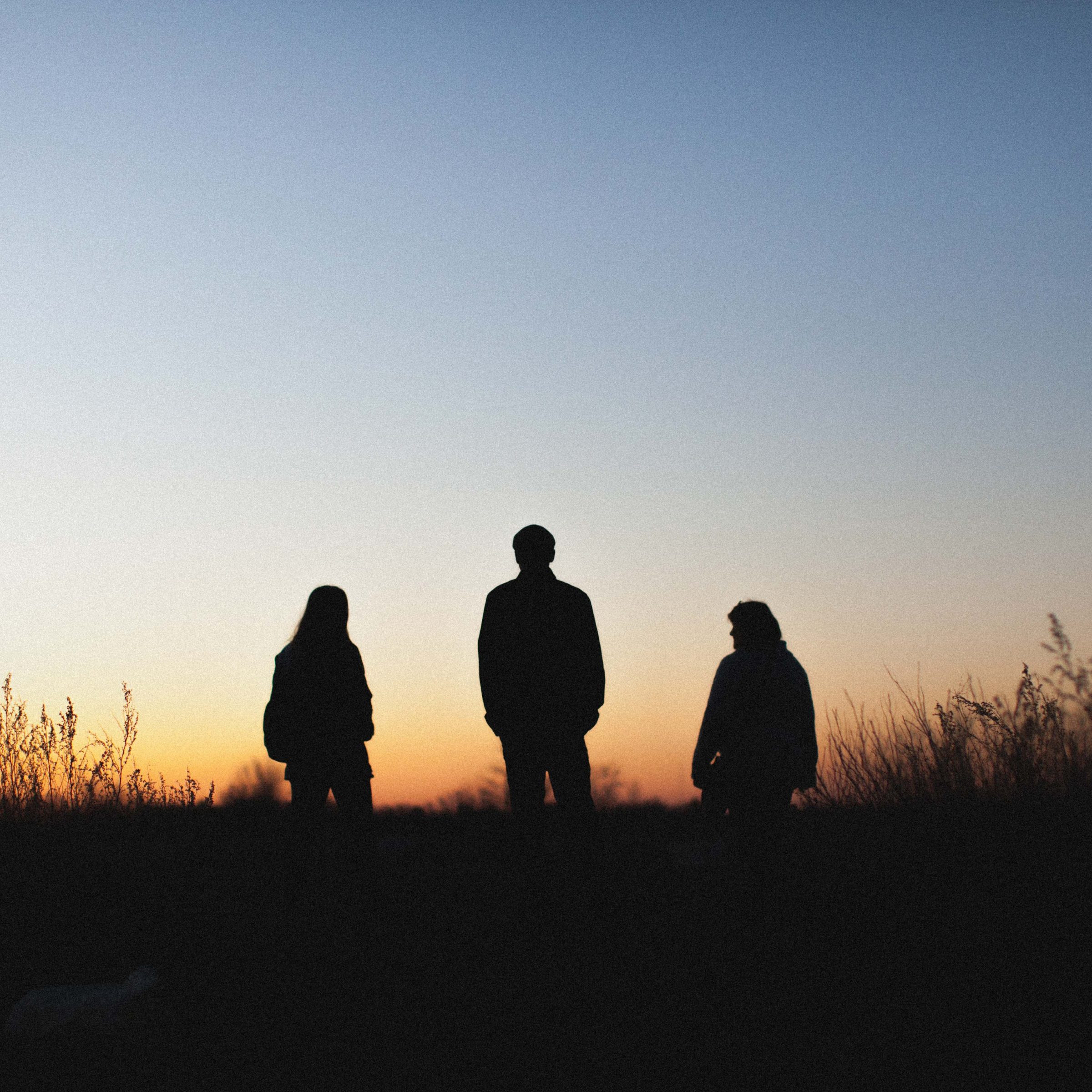 Three adult silhouettes stand in tall prairie grass watching the sunrise.