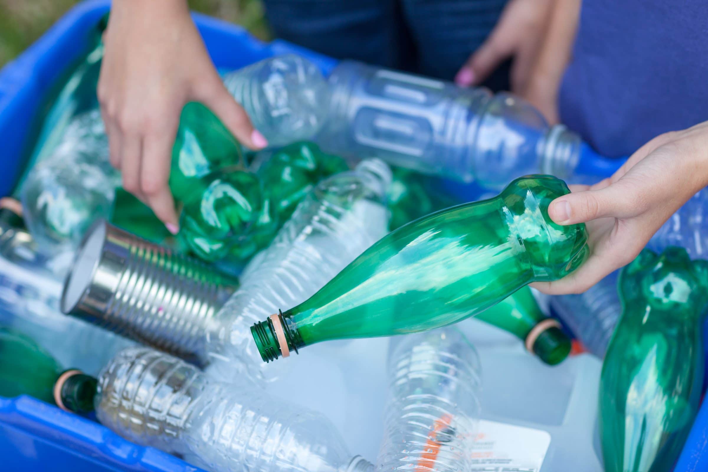 Two sets of hands organize plastic water bottles in a blue recycling bin.