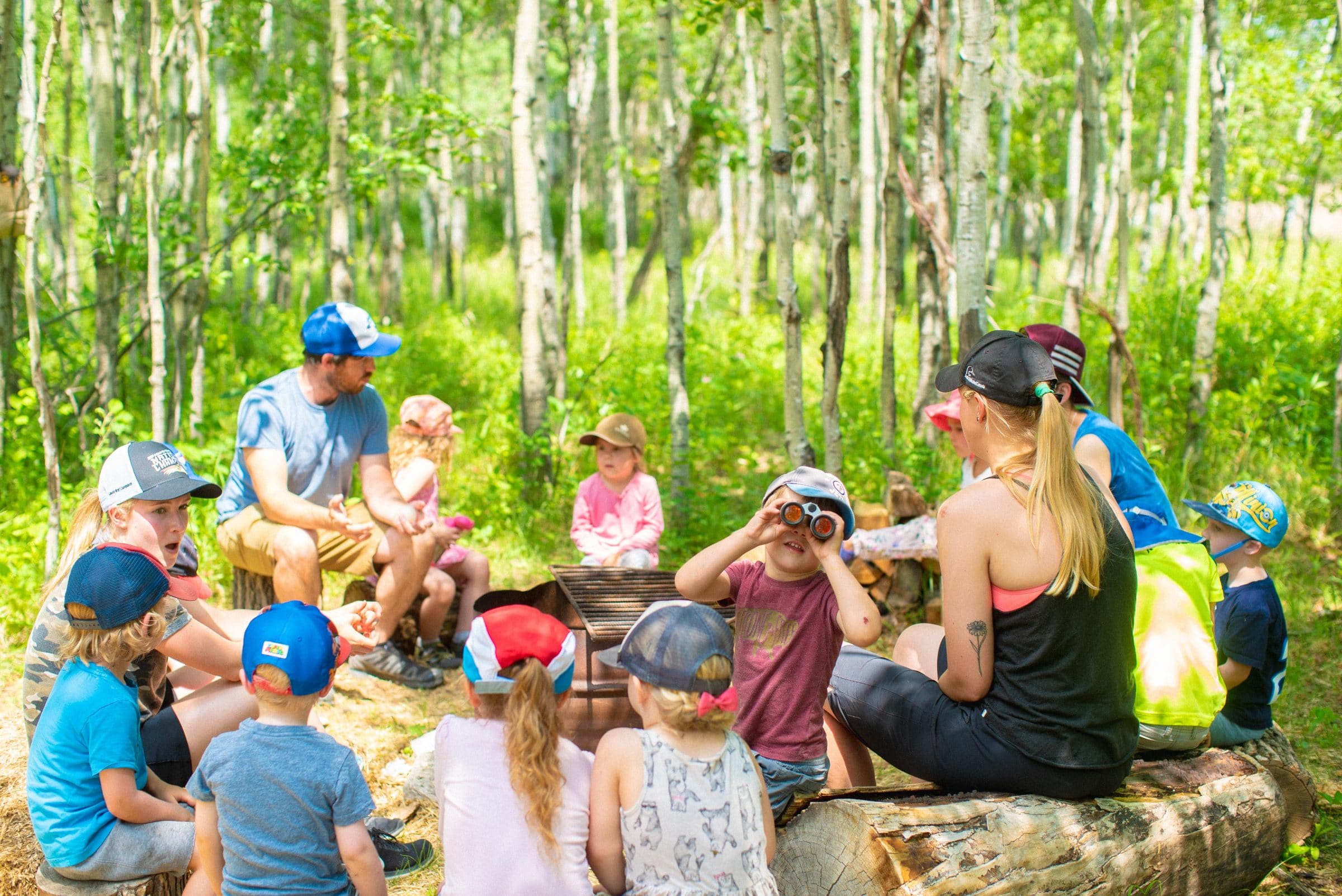 A group of kids sits on logs in the forest at Forest School.