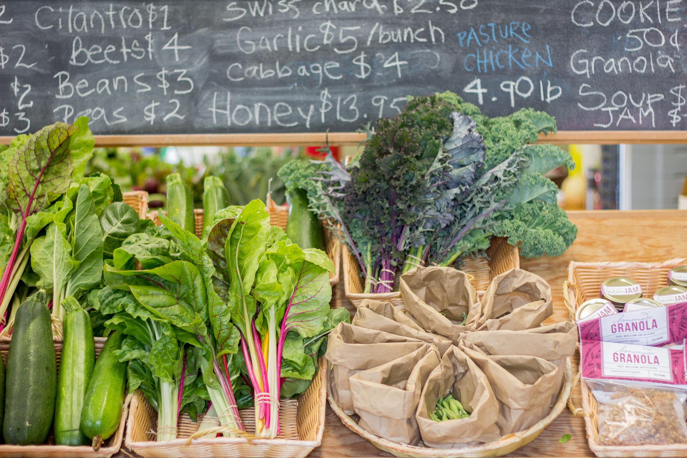 Assortment of green vegetables in baskets in front of chalkboard with price list.